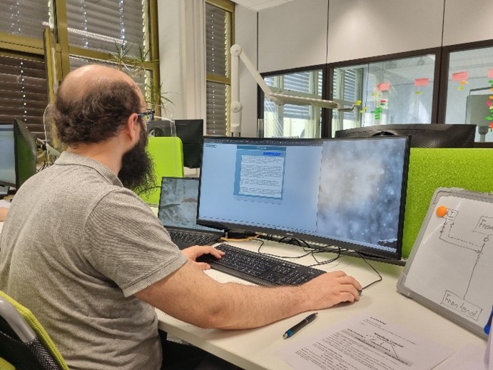 Dargestellt ist eine sitzende Person vor einem großen gebogenen Bildschirm. Auf dem Bildschirm wird ein Chatbot-Fenster angezeigt mit dem die Person interagiert