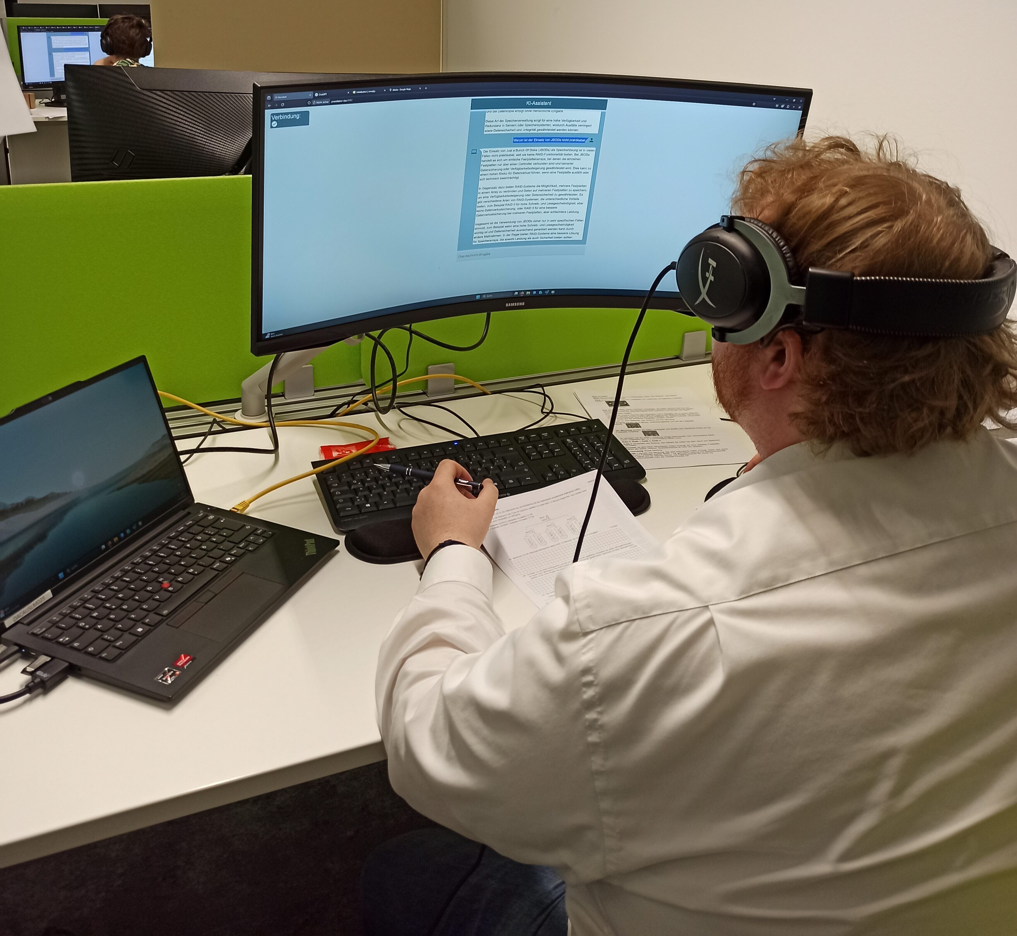 Dargestellt ist eine sitzende Person mit Headset am Arbeitsplatz vor einem großen, gebogenen Monitor. Auf dem Monitor ist eine Chatbot-Oberfläche (wie im vorherigen Bild) angezeigt. 