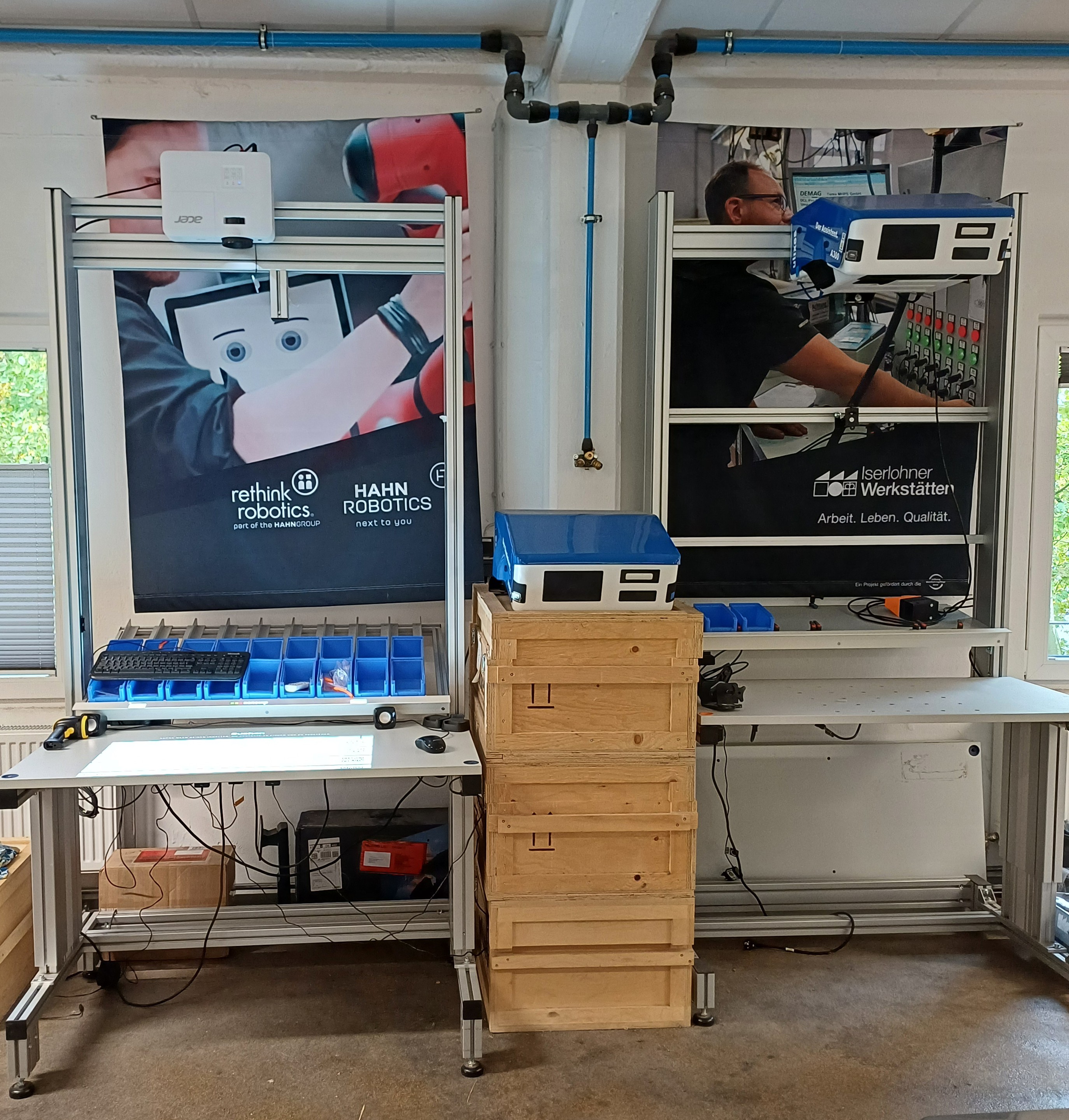Zwei nebeneinanderstehende Arbeitsstationen in einer Werkstatt, ausgestattet mit Metallrahmen, Sortierboxen, Monitoren und technischen Geräten. Im Hintergrund hängen große Poster. Auf den Tischen befinden sich Kabel und Werkzeuge, daneben mehrere Holzkisten.