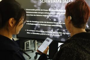 Zwei Personen schauen gemeinsam auf ein Handy und einen einen großen Bildschirm. Auf dem großen Bildschirm ist verschwommen ein Text mit der Überschrift Schwermetalle zu sehen.
