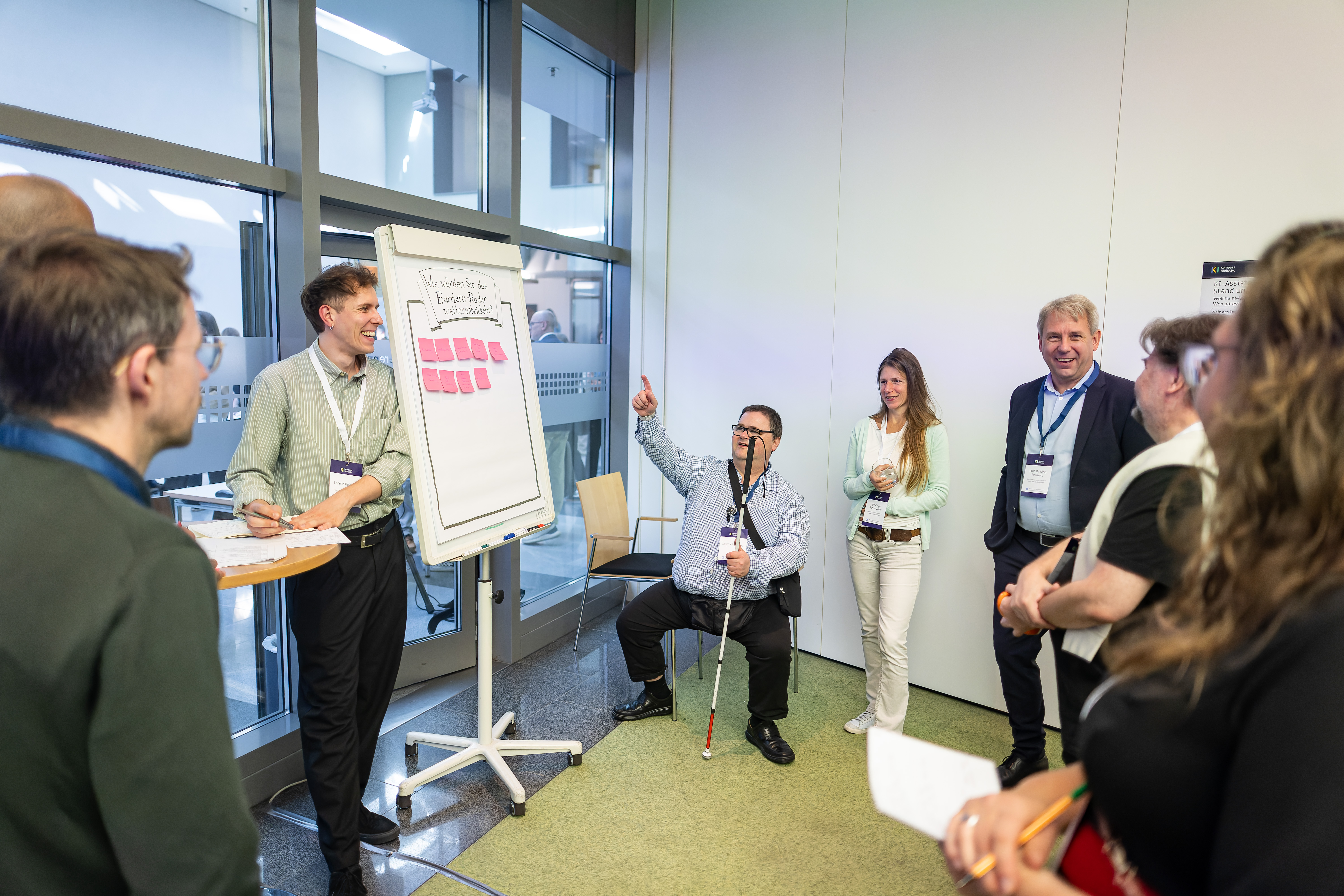 Foto, das eine kleine Gruppe lächelnder Menschen vor einem Flipchart zeigt. Eine Person schreibt mit, eine andere Person meldet sich.