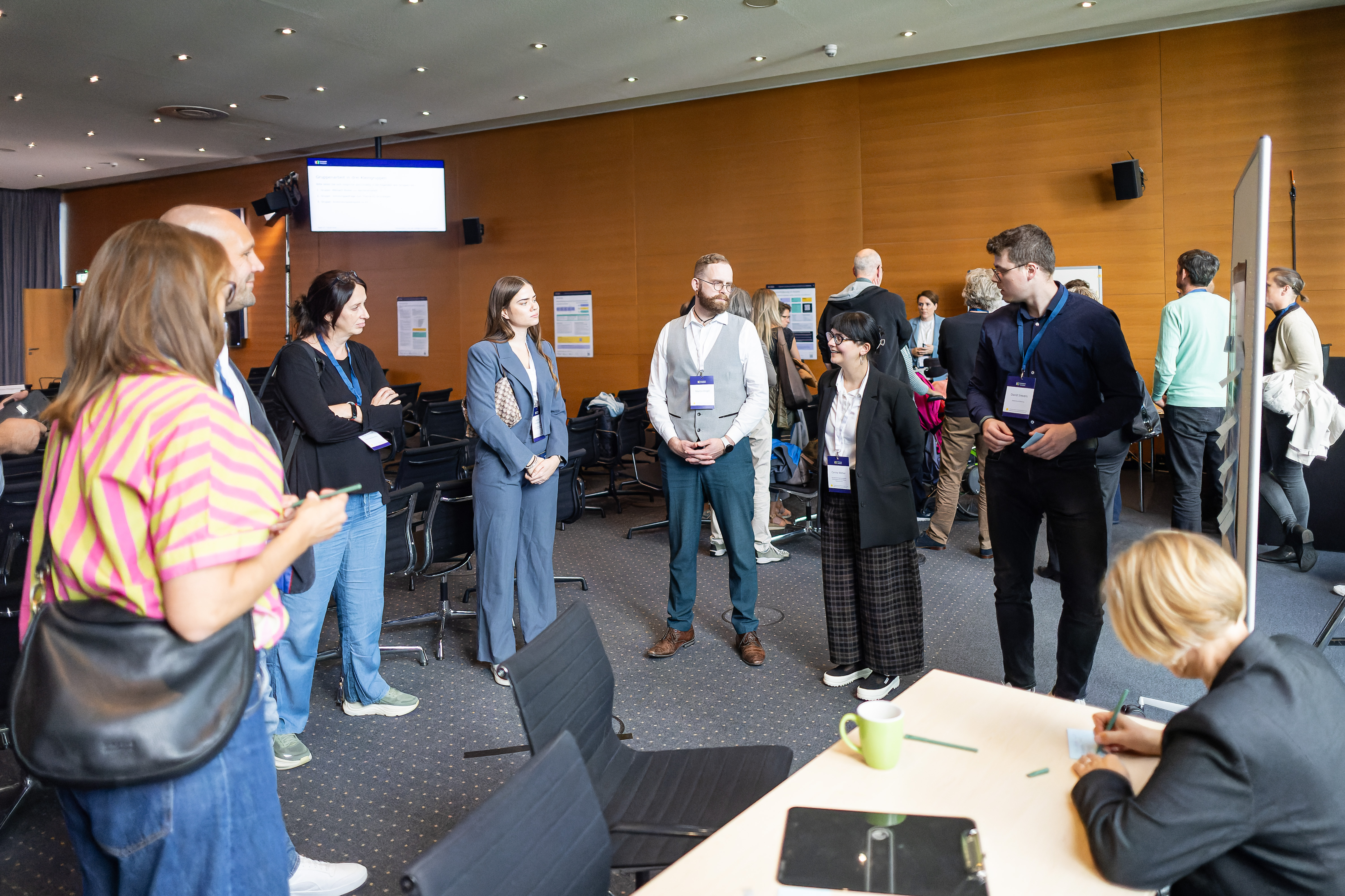 Foto einer kleinen Gruppe von Menschen, die im Halbkreis um ein Flipchart steht. Eine Person spricht, die anderen hören zu. Im Vordergrund schreibt eine Person auf ein Kärtchen.