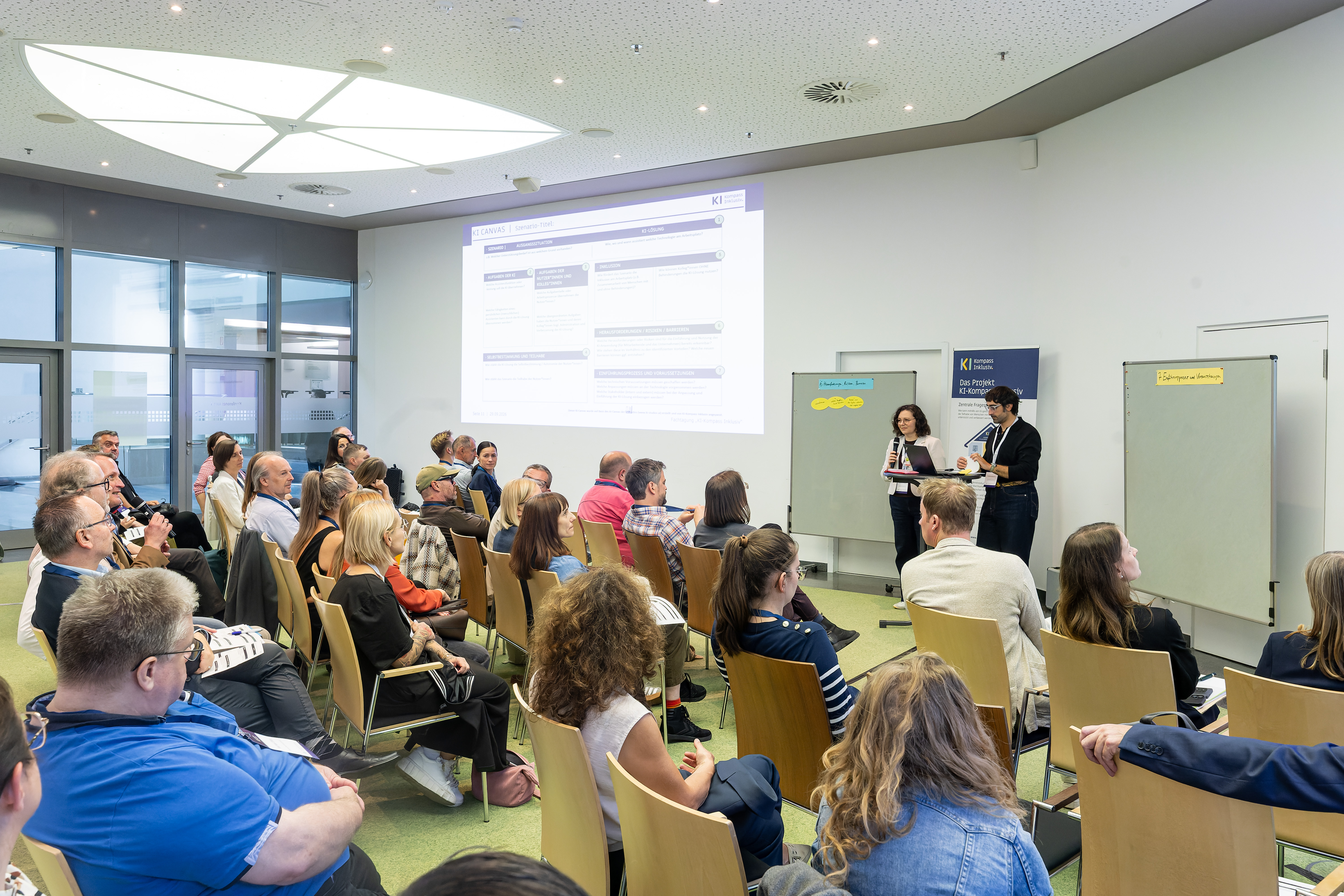 Foto vom Seminarraum. Die Teilnehmenden sitzen in Stuhlreihen in Richtung von zwei Vortragenden. Hinter ihnen eine Powerpoint Präsentation und zwei Flipcharts