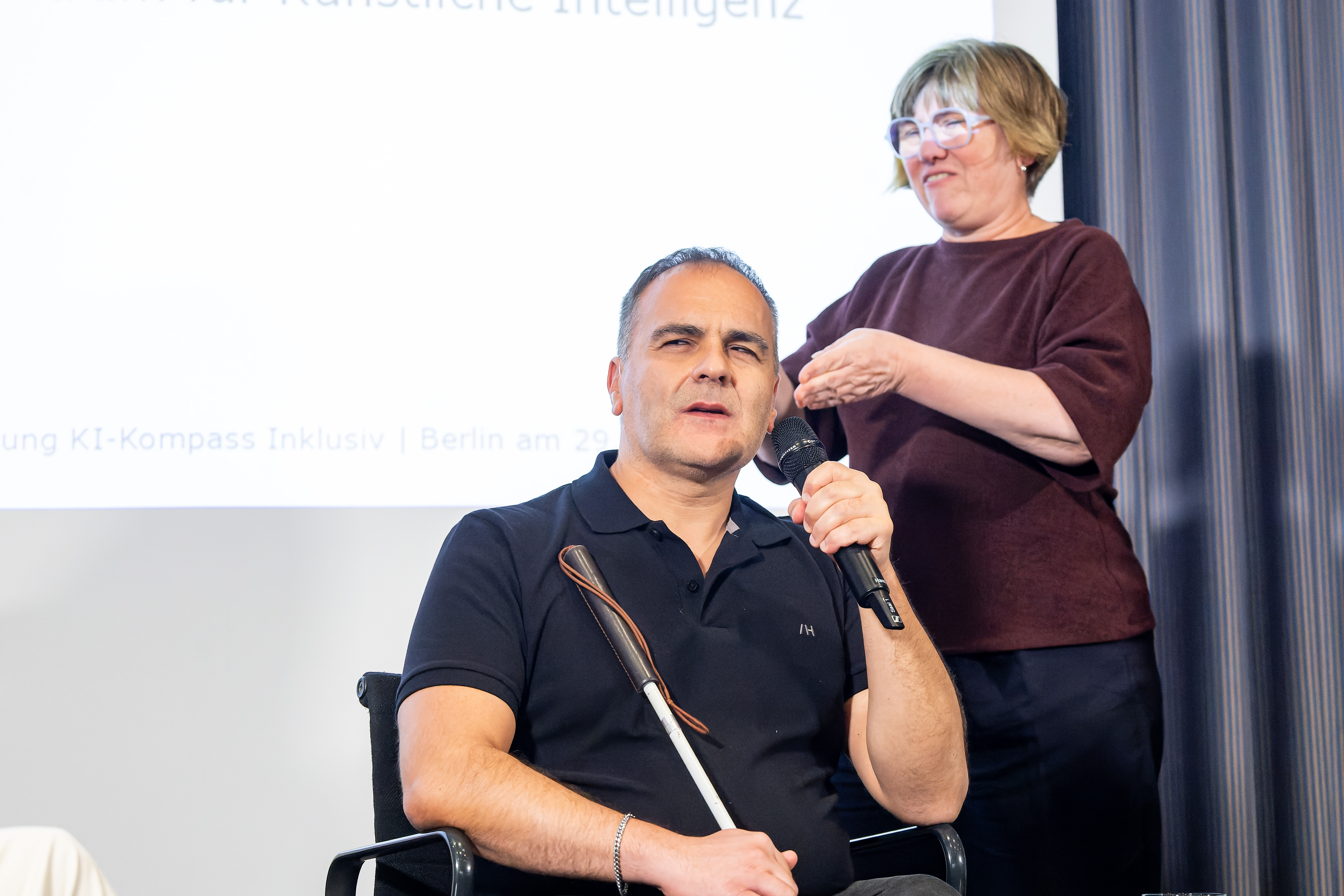 Foto von Hakan Calim, der gerade in ein Mikrofon spricht. Hinter ihm steht eine Gebärdensprach-Dolmetscherin